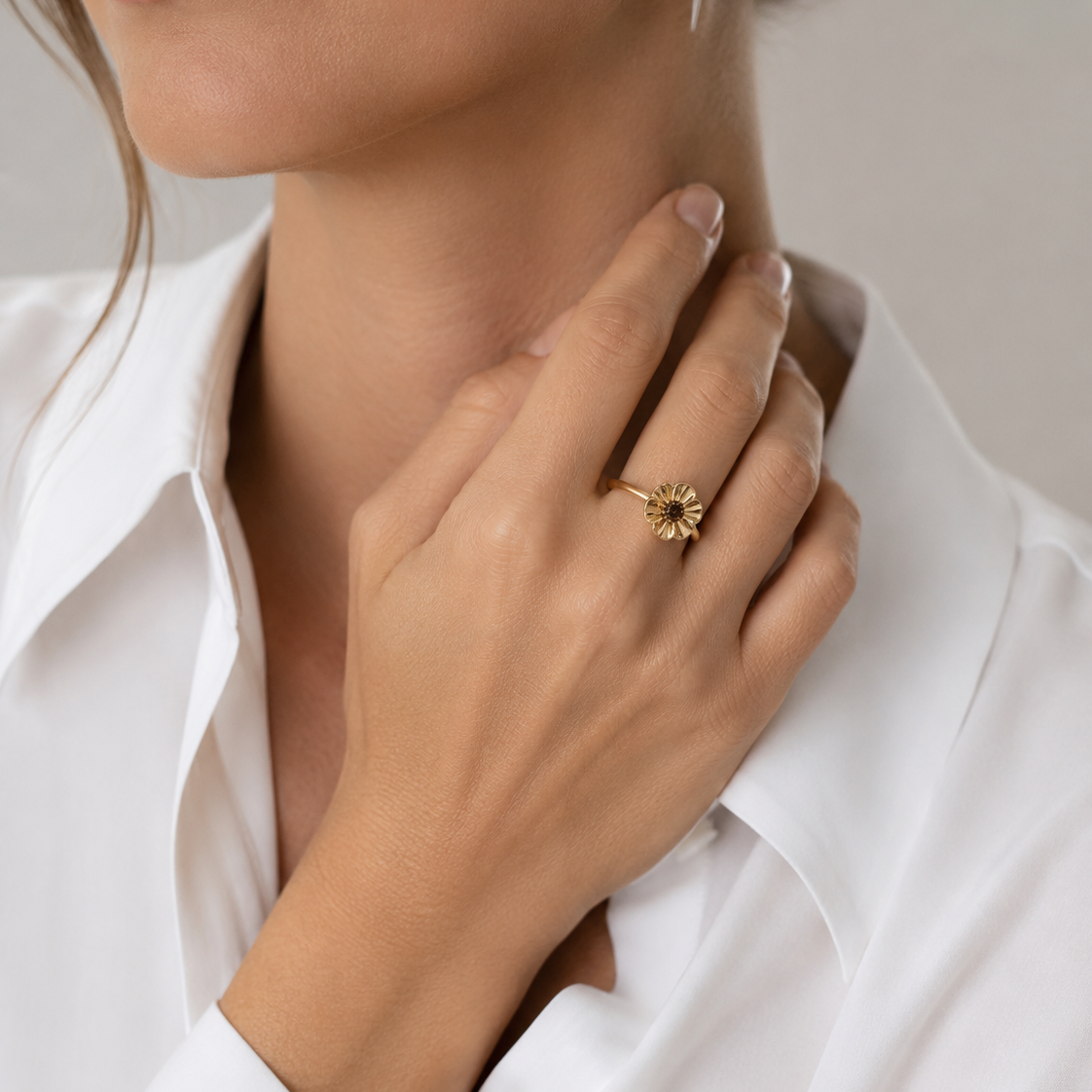 Close-up of a hand wearing a gold ring with a sunflower design, touching the collarbone against a neutral background.
